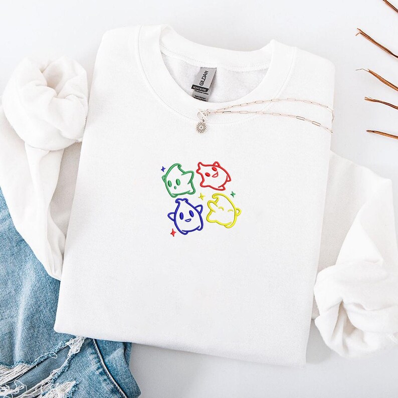 May include: White crewneck sweatshirt with a colorful embroidered design of four cartoon characters. Each character is a different color: green, red, blue, and yellow. A silver necklace with a small pendant is also visible.