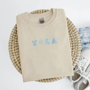 May include: Beige crewneck sweatshirt with light blue embroidered design of a glove, snowflake, dress, and castle. The sweatshirt is folded on a woven basket with denim shorts and white sneakers in the background.