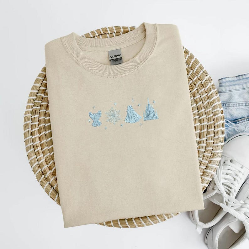May include: Beige crewneck sweatshirt with light blue embroidered design of a glove, snowflake, dress, and castle. The sweatshirt is folded on a woven basket with denim shorts and white sneakers in the background.