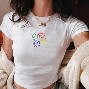 May include: White t-shirt with a crew neck and short sleeves. The shirt features a colorful embroidered design of four cartoon ghosts in green, red, blue, and yellow. The design is centered on the chest.