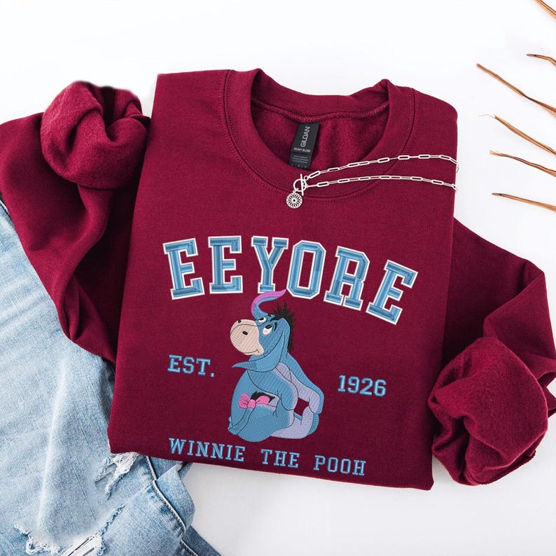 May include: A maroon sweatshirt featuring the word "Eeyore" in blue lettering, with an image of Eeyore and the text "EST. 1926 WINNIE THE POOH." A silver necklace is draped across the sweatshirt. The sleeves are folded.