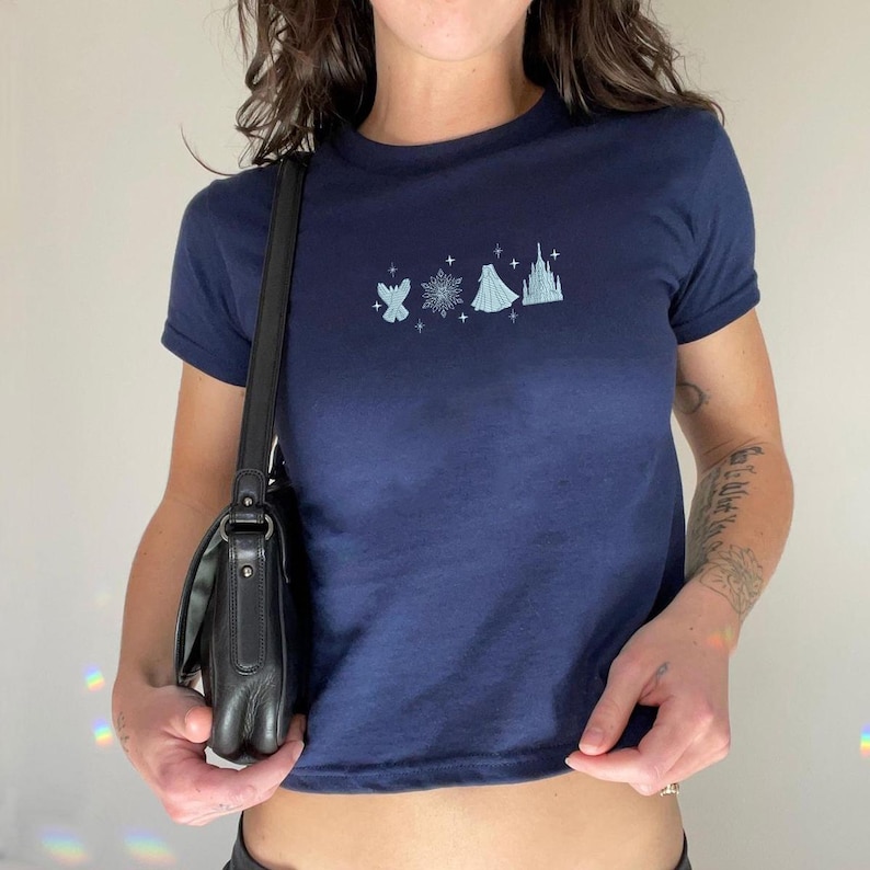 May include: Navy blue crop top with a light blue graphic design featuring a snowflake, castle, and other elements. The shirt has short sleeves and a crew neck. The model is wearing a black purse.