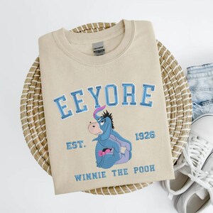 May include: Beige crewneck sweatshirt with the word "Eeyore" in blue lettering, along with an image of Eeyore from Winnie the Pooh. The sweatshirt also has the text "EST. 1926" and "Winnie the Pooh" in blue.