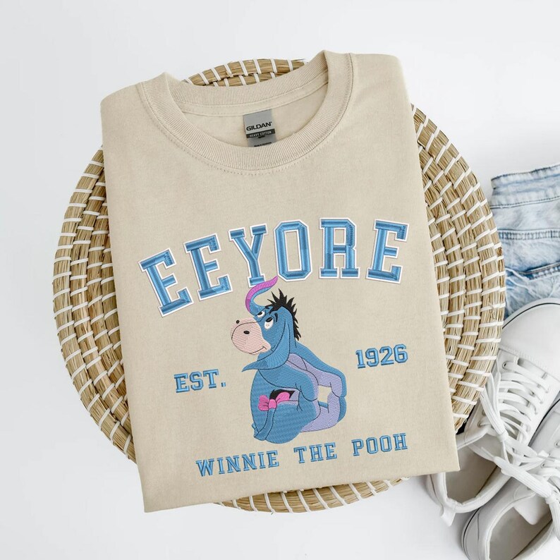 May include: Beige crewneck sweatshirt with the word "Eeyore" in blue lettering, along with an image of Eeyore from Winnie the Pooh. The sweatshirt also has the text "EST. 1926" and "Winnie the Pooh" in blue.