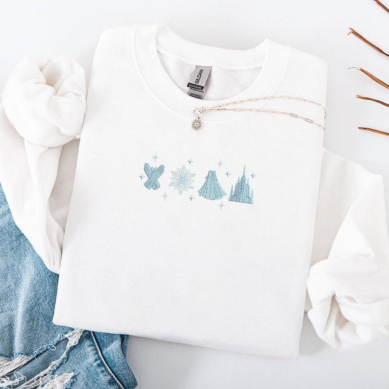 May include: White crewneck sweatshirt with light blue embroidered design featuring a snowflake, gloves, dress, and castle. A silver necklace with a sun pendant rests on the sweatshirt. The sweatshirt is folded on top of blue jeans.