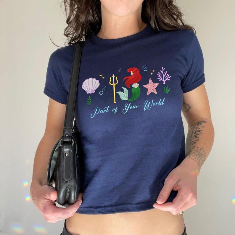 May include: A navy blue crop top with a colourful graphic of a mermaid, seashell, starfish, and coral. The text "Part of Your World" is printed in a light blue script. The shirt has short sleeves.
