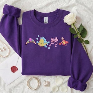 May include: A purple sweatshirt with embroidered images of a tiara, a yellow and blue fish, a pink bow, and a red crab. The sweatshirt has long sleeves with rolled cuffs. A white rose and pearl necklace are also visible.
