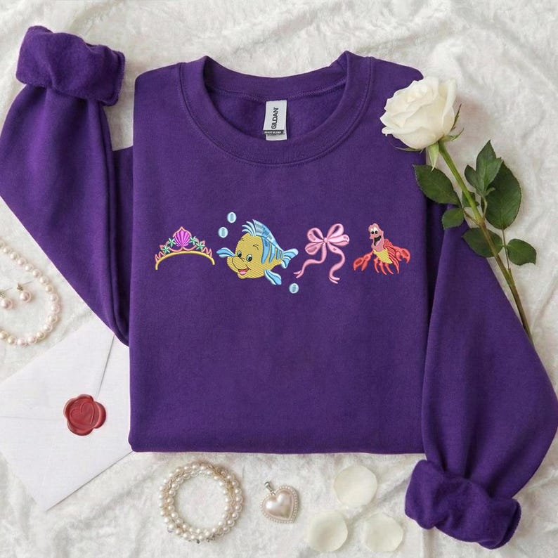 May include: A purple sweatshirt with embroidered images of a tiara, a yellow and blue fish, a pink bow, and a red crab. The sweatshirt has long sleeves with rolled cuffs. A white rose and pearl necklace are also visible.