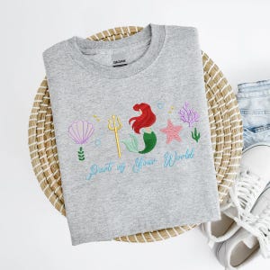 May include: A grey crew neck sweatshirt with an embroidered design. It features a mermaid with red hair and a green tail, a golden trident, and the text "Part of Your World" in light blue. The design also includes a seashell, starfish, and coral.
