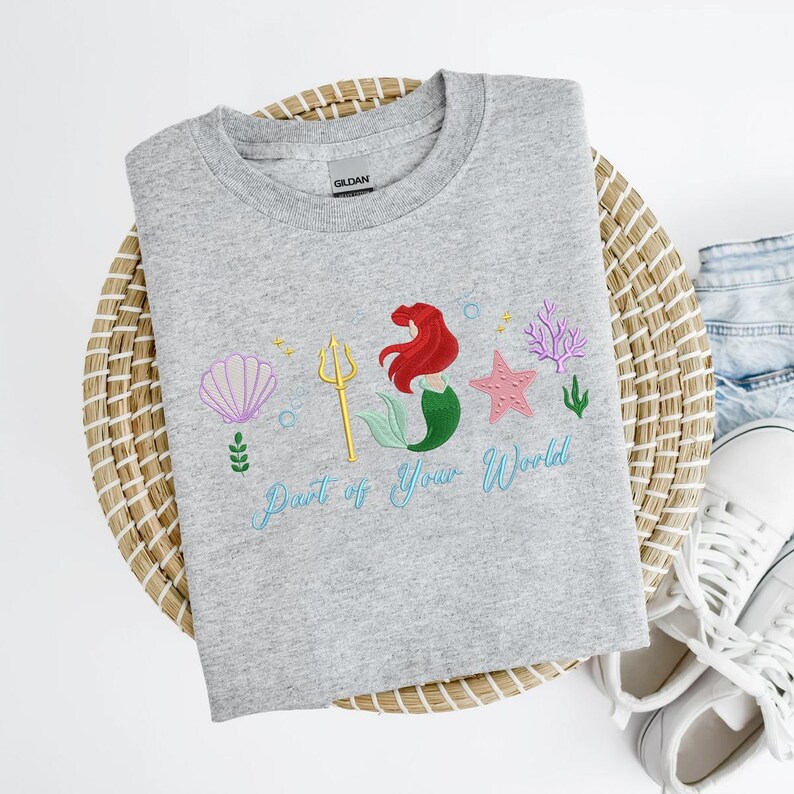 May include: A grey crew neck sweatshirt with an embroidered design. It features a mermaid with red hair and a green tail, a golden trident, and the text "Part of Your World" in light blue. The design also includes a seashell, starfish, and coral.