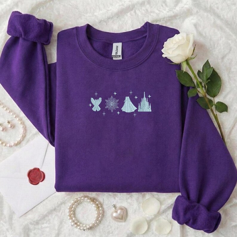 May include: A purple sweatshirt with a crew neck and cuffed sleeves. The front features a light blue embroidered design of a fairy, snowflake, dress, and castle. A white rose and jewelry are in the background.