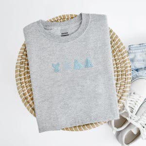 May include: A heather gray crewneck sweatshirt with light blue embroidered designs of a cactus, a star, a sail, and a building. The sweatshirt is on a woven basket with white sneakers and denim jeans.