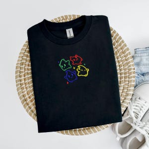 May include: A black t-shirt featuring a colorful embroidered design of four cartoon characters. The characters are outlined in green, red, blue, and yellow, with matching colored accents. The shirt is displayed on a woven basket.