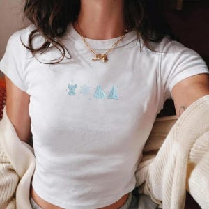 May include: White t-shirt with short sleeves featuring light blue embroidered designs of a bow, snowflake, and two structures. The shirt is made of a soft material and is a casual top.