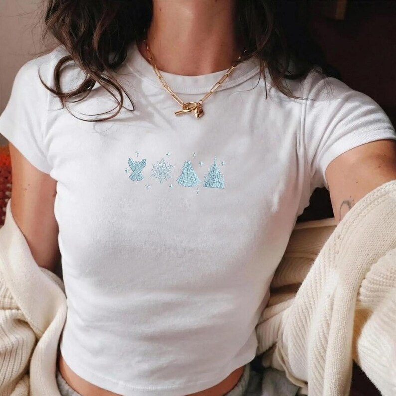 May include: White t-shirt with short sleeves featuring light blue embroidered designs of a bow, snowflake, and two structures. The shirt is made of a soft material and is a casual top.