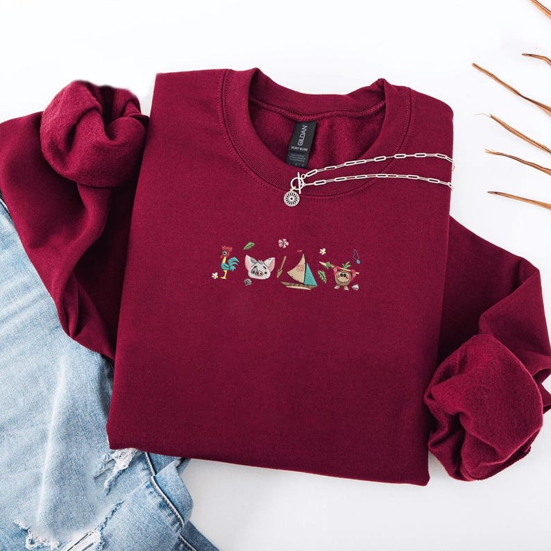 May include: A maroon crewneck sweatshirt with embroidered cartoon characters and a silver necklace. The embroidered design includes a rooster, a cat, a sailboat, and a character with a leaf skirt. The sweatshirt is folded on top of blue jeans.