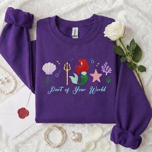 May include: A purple sweatshirt featuring a mermaid, seashell, starfish, and trident graphic. The text "Part of Your World" is below the graphic. The sweatshirt has long sleeves with cuffed wrists.