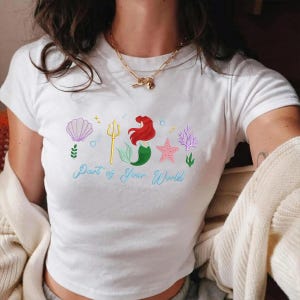 May include: White t-shirt with a colourful graphic featuring a mermaid, seashell, starfish, and coral. The text "Part of Your World" is written in light blue script below the graphic. The design includes a golden trident.