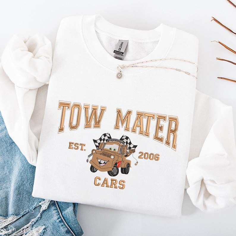 Puede incluir: Una sudadera blanca con un dise&ntilde;o dorado de Tow Mater de la pel&iacute;cula Cars. El dise&ntilde;o incluye el texto "TOW MATER", "EST. 2006" y "CARS". Tambi&eacute;n se ve un collar plateado con un peque&ntilde;o colgante. La sudadera est&aacute; doblada sobre mezclilla.