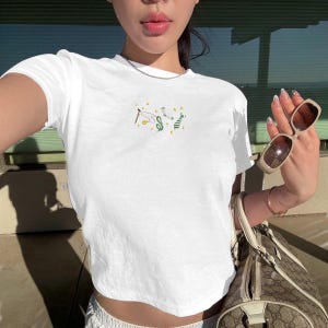 May include: White crop top with a small embroidered design featuring a dollar sign, golf club, and other symbols. The shirt is short-sleeved and has a crew neck. The design is in shades of green, yellow, and brown.