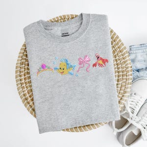 May include: A light gray sweatshirt featuring embroidered images of a tiara, a yellow fish, a pink bow, and a red crab. The sweatshirt is laid on a woven basket with a pair of white sneakers and blue jeans.