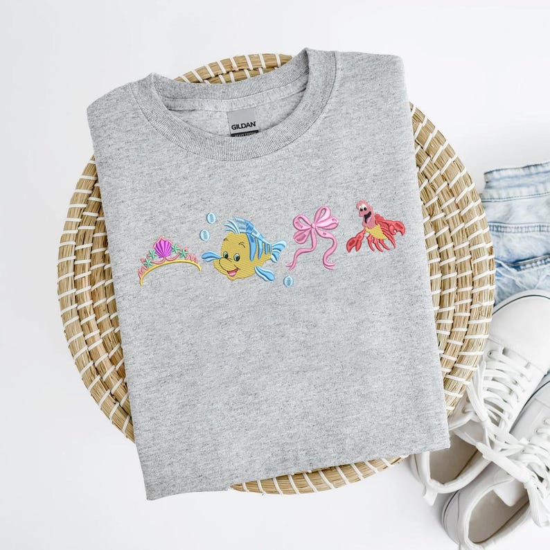 May include: A light gray sweatshirt featuring embroidered images of a tiara, a yellow fish, a pink bow, and a red crab. The sweatshirt is laid on a woven basket with a pair of white sneakers and blue jeans.
