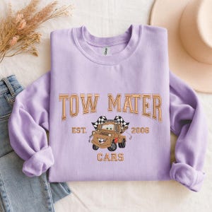 Puede incluir: Una sudadera lila con la inscripci&oacute;n "TOW MATER" en letras doradas. Debajo, una ilustraci&oacute;n de Mater de la pel&iacute;cula Cars, con el texto "EST. 2006 CARS". La sudadera tiene mangas largas y cuello redondo.