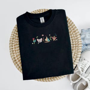 May include: Black crew neck t-shirt with embroidered cartoon characters including a rooster, pig, sailboat, and monkey. The shirt is displayed on a woven basket with white sneakers and denim jeans in the background.