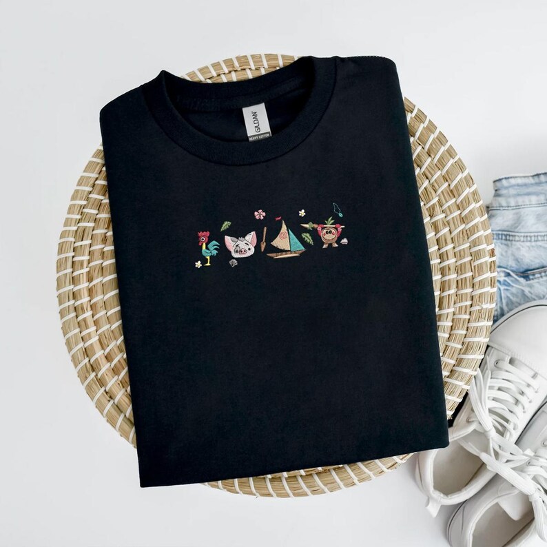 May include: Black crew neck t-shirt with embroidered cartoon characters including a rooster, pig, sailboat, and monkey. The shirt is displayed on a woven basket with white sneakers and denim jeans in the background.