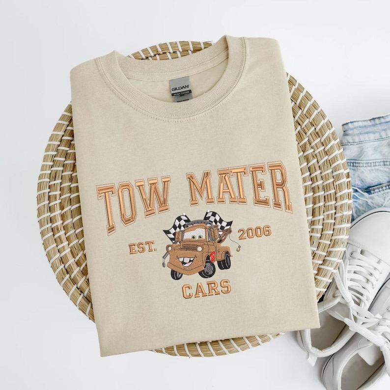 Puede incluir: Sudadera beige con un dise&ntilde;o bordado de Mate de la pel&iacute;cula Cars. El dise&ntilde;o incluye el texto "TOW MATER" encima del personaje, con "EST. 2006" y "CARS" debajo. La sudadera est&aacute; doblada sobre una estera tejida.