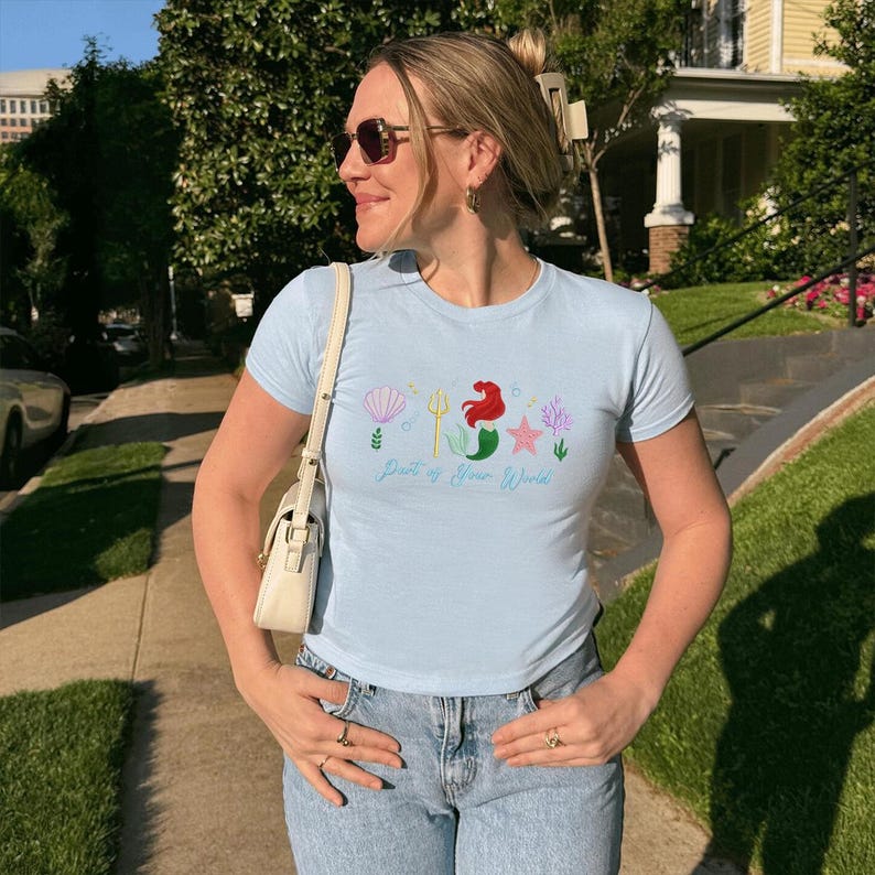 May include: Pale blue t-shirt featuring a mermaid graphic and the text "Part of Your World". The design includes a seashell, trident, starfish, and seaweed. The shirt is worn with blue jeans.