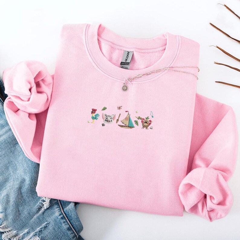 May include: A light pink sweatshirt with embroidered cartoon characters and a sailboat design. A silver necklace with a sun pendant is on the sweatshirt. The sweatshirt is on a white surface with a pair of blue jeans.