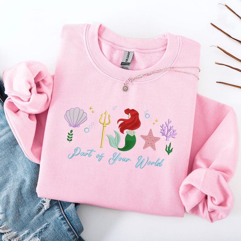May include: A pale pink sweatshirt with an embroidered design of a mermaid, seashell, starfish, and seaweed. The words "Part of Your World" are written in a script font below the design. A silver necklace with a small pendant is on the sweatshirt.