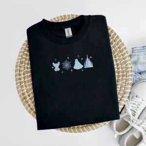 May include: Black crew neck t-shirt with light blue embroidered design featuring a snowflake, a dress, a castle, and other elements. The shirt is displayed on a woven basket with white sneakers and denim jeans in the background.