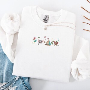 May include: White crewneck sweatshirt with embroidered cartoon characters, including a rooster, pig, sailboat, and bear. A silver necklace with a pendant is draped across the sweatshirt. The sweatshirt is folded on top of blue denim jeans.