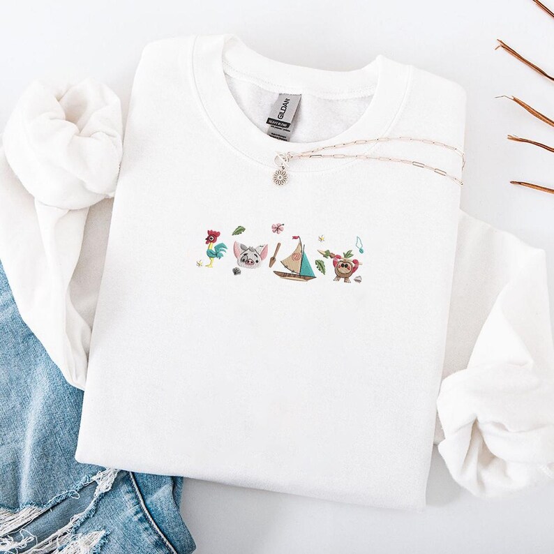 May include: White crewneck sweatshirt with embroidered cartoon characters, including a rooster, pig, sailboat, and bear. A silver necklace with a pendant is draped across the sweatshirt. The sweatshirt is folded on top of blue denim jeans.