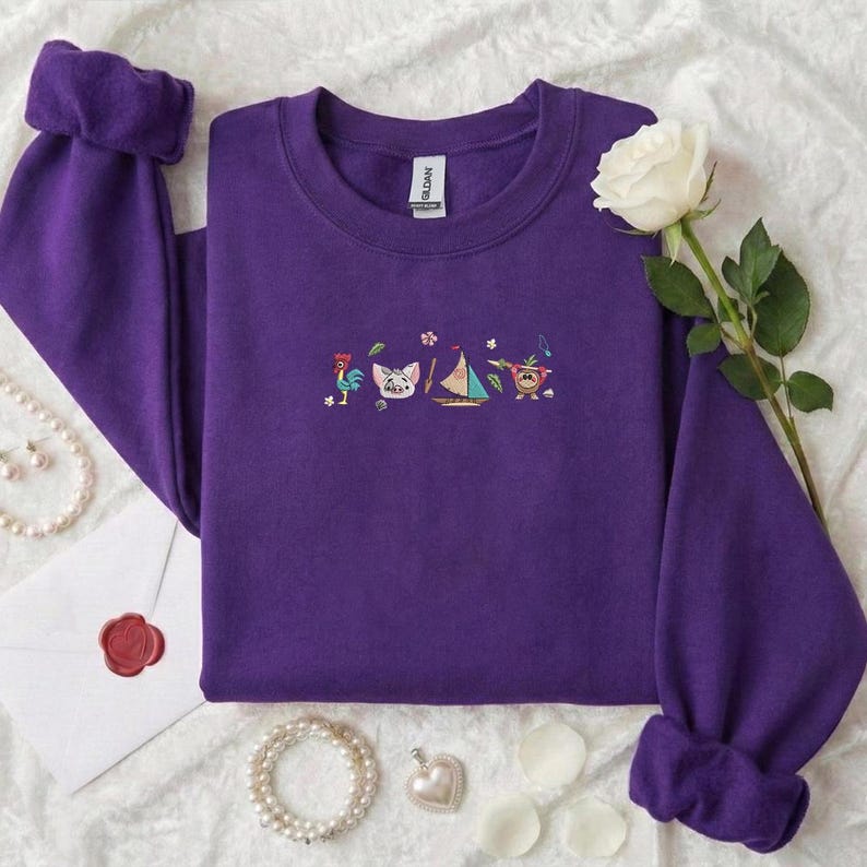 May include: A purple sweatshirt with embroidered cartoon characters. The design includes a rooster, pig, sailboat, and a character with musical notes. The sleeves are cuffed. A white rose and jewelry are in the background.