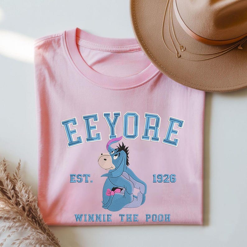May include: A light pink t-shirt featuring a graphic of Eeyore from Winnie the Pooh. The design includes the name "Eeyore" in blue block letters, the text "EST. 1926", and the words "Winnie the Pooh". A brown hat is in the background.