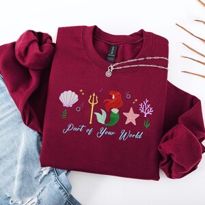 May include: Burgundy sweatshirt with an embroidered mermaid design. The design includes a seashell, trident, mermaid, starfish, and coral. The text "Part of Your World" is embroidered below the design. A silver necklace is draped on the sweatshirt.
