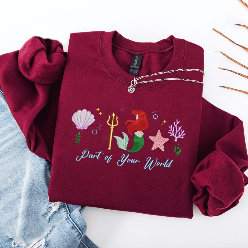 May include: Burgundy sweatshirt with an embroidered mermaid design. The design includes a seashell, trident, mermaid, starfish, and coral. The text "Part of Your World" is embroidered below the design. A silver necklace is draped on the sweatshirt.