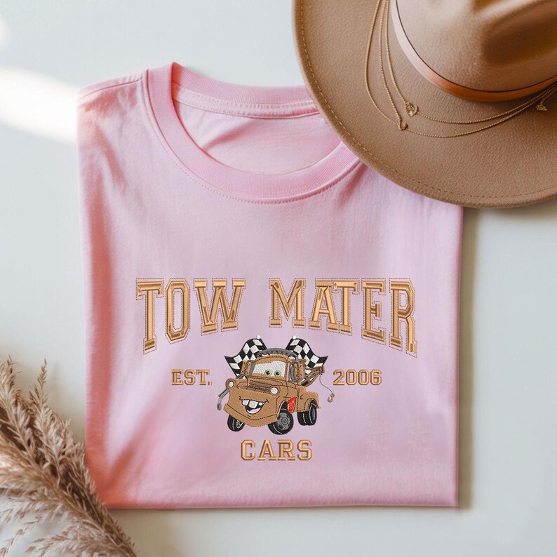 Puede incluir: Camiseta rosa claro con la inscripci&oacute;n "TOW MATER" en letras doradas y una imagen de Mater de la pel&iacute;cula Cars. La camiseta tambi&eacute;n incluye "EST. 2006" y la palabra "CARS". Un sombrero beige est&aacute; en el fondo.
