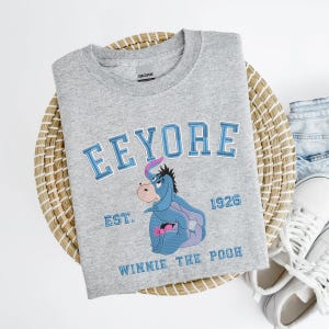 May include: A gray crew neck sweatshirt featuring the word "Eeyore" in blue lettering above an embroidered image of Eeyore from Winnie the Pooh. The sweatshirt also includes the text "EST. 1926" and "Winnie the Pooh" in blue.