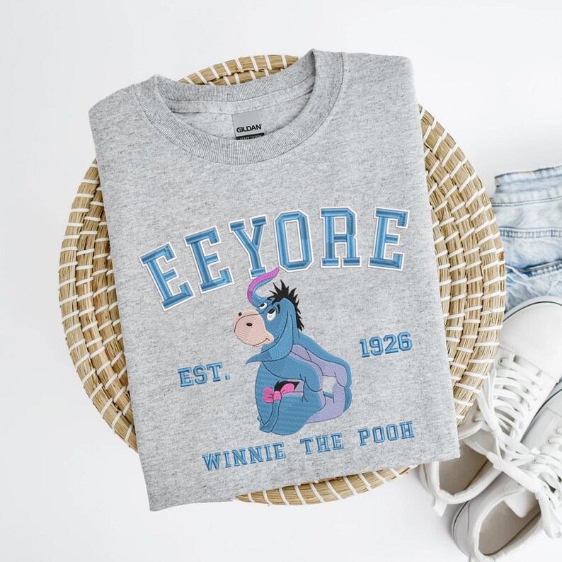 May include: A gray crew neck sweatshirt featuring the word "Eeyore" in blue lettering above an embroidered image of Eeyore from Winnie the Pooh. The sweatshirt also includes the text "EST. 1926" and "Winnie the Pooh" in blue.