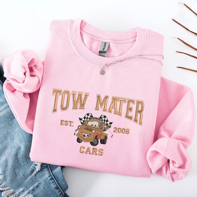 Puede incluir: Sudadera rosa claro con la inscripci&oacute;n "TOW MATER" y una ilustraci&oacute;n de Mate de Cars. El dise&ntilde;o incluye "EST. 2006" y "CARS". Un collar plateado est&aacute; sobre la sudadera.