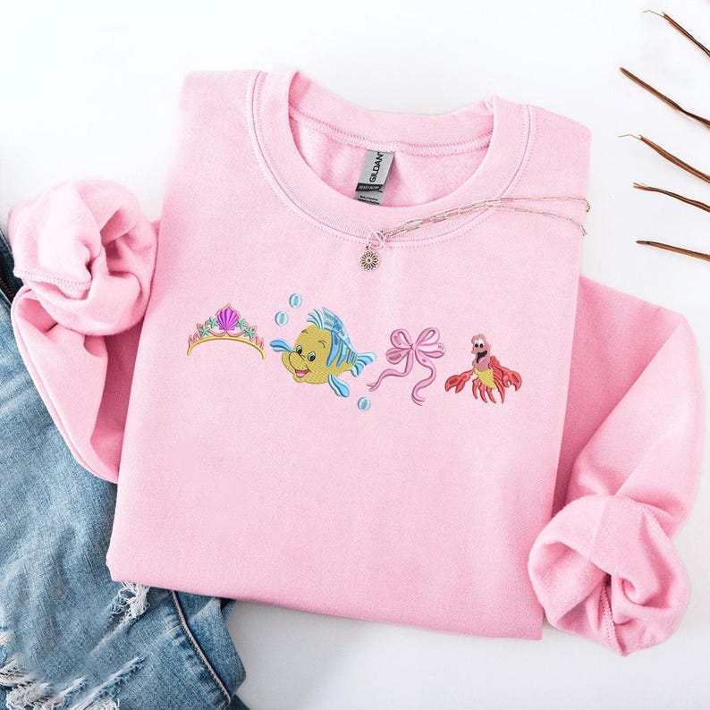 May include: A light pink sweatshirt featuring embroidered images of a tiara, a yellow and blue fish, a pink bow, and a red crab. A silver necklace with a sun pendant is draped across the sweatshirt. The sweatshirt is folded on top of a pair of blue jeans.