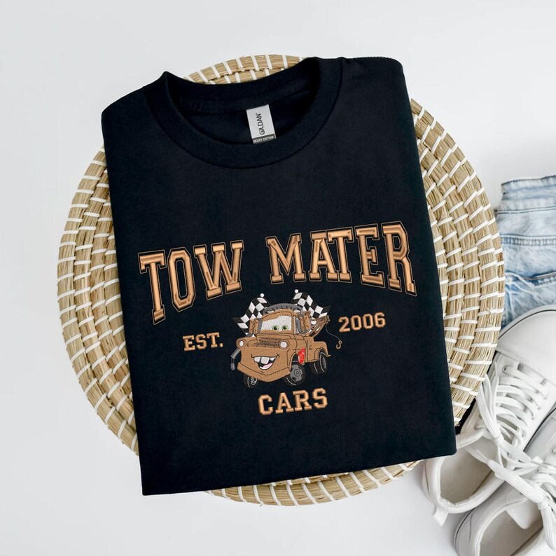 Puede incluir: Camiseta negra con un dise&ntilde;o de Mate de la pel&iacute;cula Cars. El dise&ntilde;o incluye el texto "TOW MATER" encima del personaje, con "EST. 2006" y "CARS" debajo. La camiseta se muestra sobre una cesta tejida.