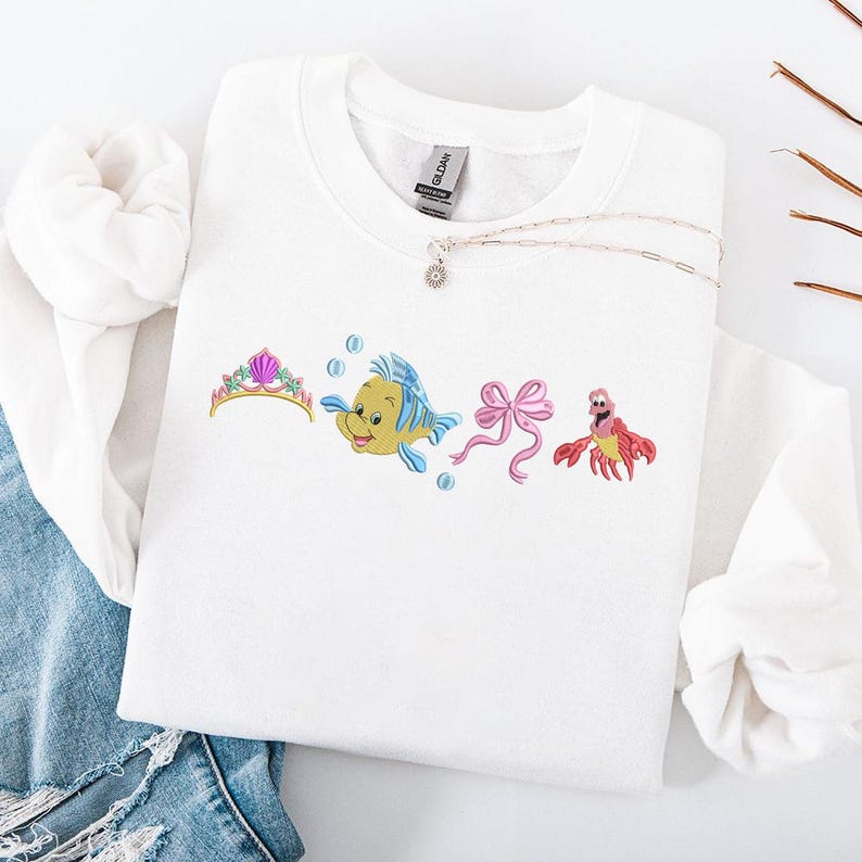 May include: White sweatshirt featuring embroidered images of a tiara, a yellow and blue fish, a pink bow, and a red crab. A silver necklace with a pendant is also visible. The sweatshirt is folded on top of a pair of blue jeans.