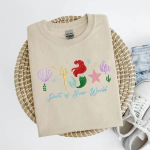 May include: Beige sweatshirt with an embroidered design featuring a mermaid, seashell, starfish, and coral. The text "Part of Your World" is embroidered in a light blue script. The sweatshirt is on a woven basket.