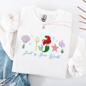 May include: White sweatshirt with an embroidered design of a mermaid, seashell, trident, starfish, and coral. The text "Part of Your World" is written in teal script. A silver necklace with a pendant is also visible.