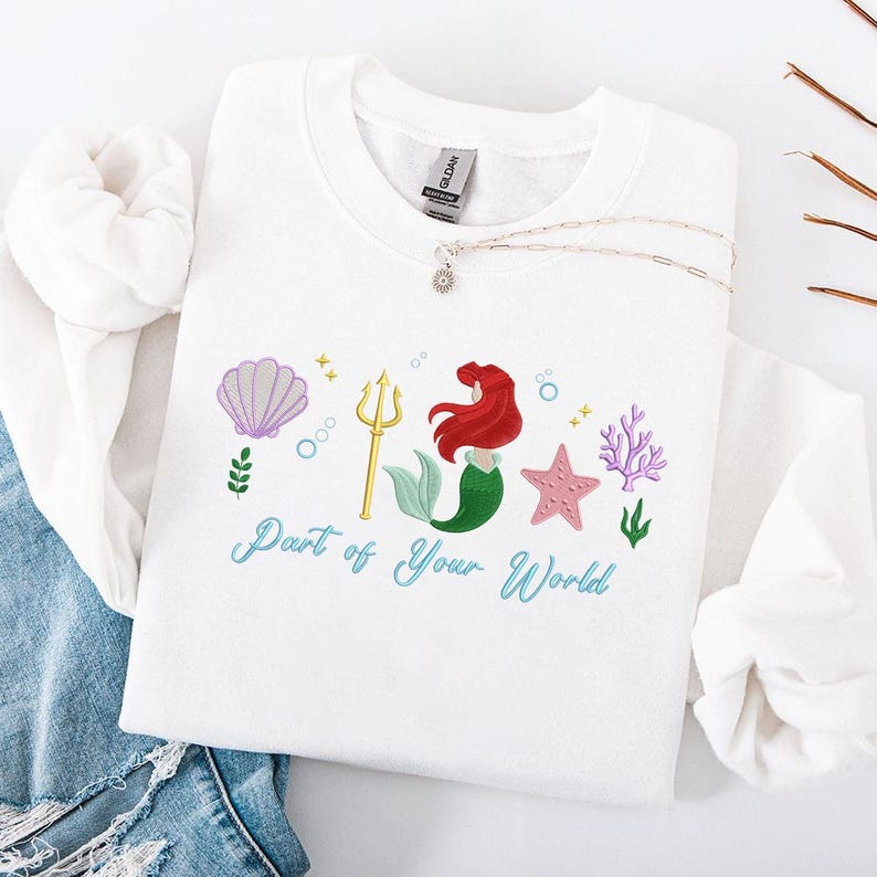 May include: White sweatshirt with an embroidered design of a mermaid, seashell, trident, starfish, and coral. The text "Part of Your World" is written in teal script. A silver necklace with a pendant is also visible.